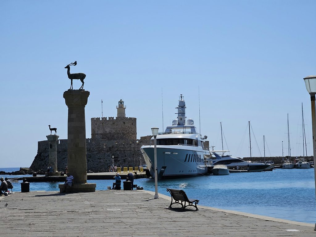 Yacht a motore ormeggiato all'ingresso di un porto con statue di cervi su colonne di pietra, Rodi