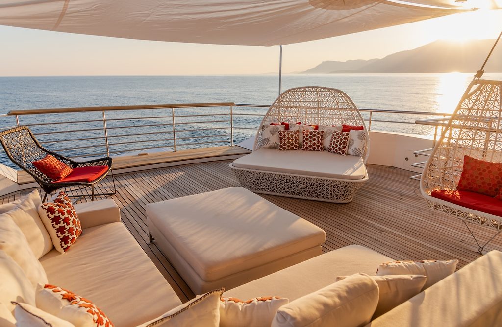 Cubierta lounge sombreada con sofás y sillones colgantes en un yate, costa al atardecer