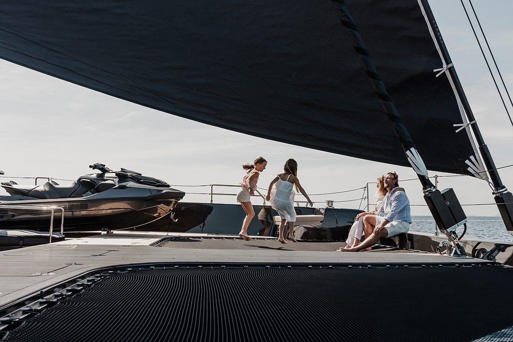 Children playing and adults relaxing on the foredeck of a sailing catamaran, jet ski stowed nearby, at sea