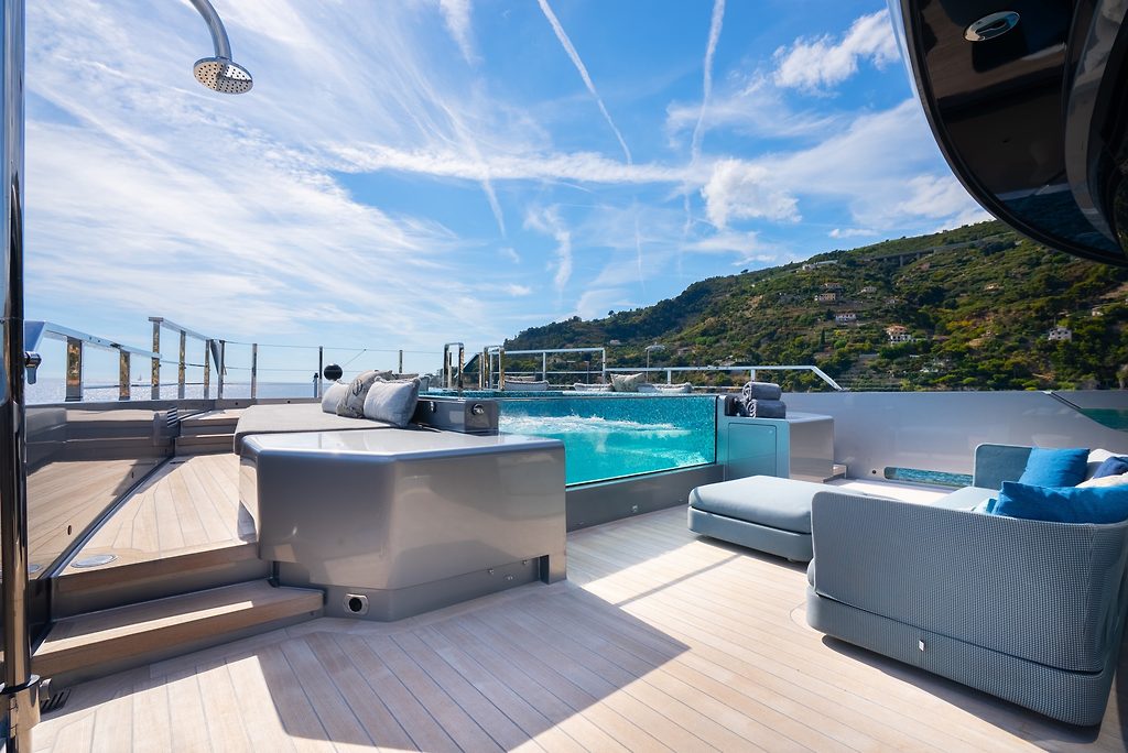 Sun deck with pool and lounge seating on a superyacht, coastal hills in the background