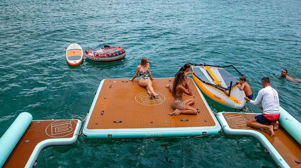Personas relajándose en una plataforma flotante inflable con tablas de paddle, nadando junto a un yate