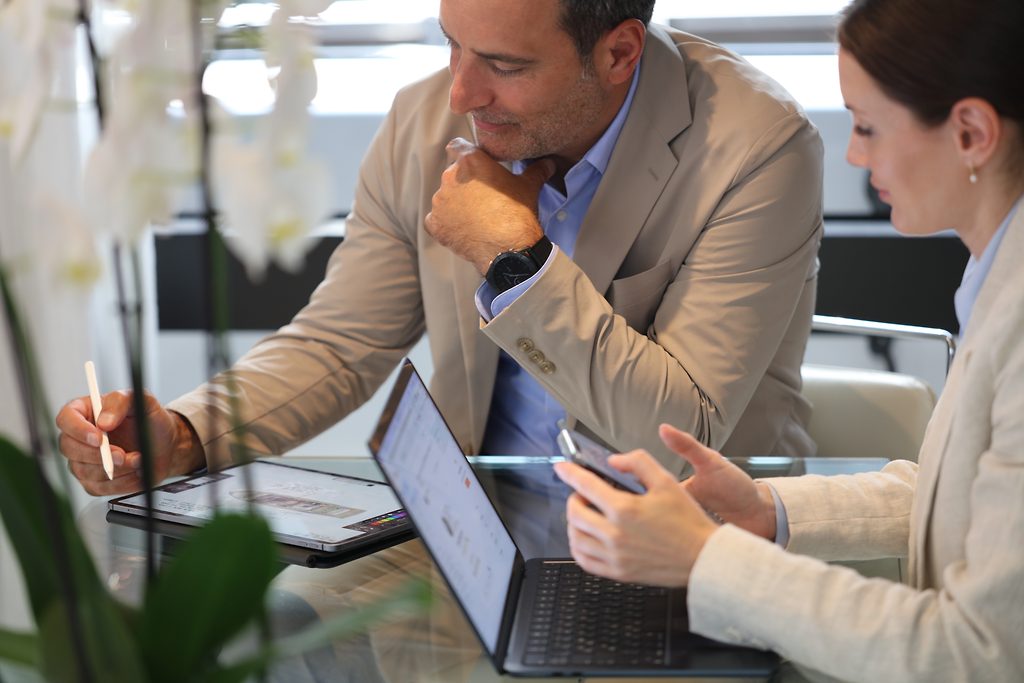 Makler und Kundin besprechen Unterlagen auf Tablet und Laptop an einem Bürotisch