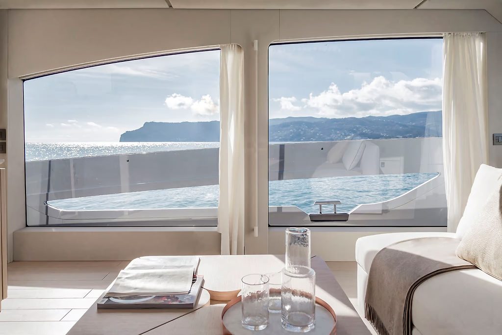 Salón de yate con grandes ventanas hacia la cubierta de popa y la costa, mesa dispuesta con vasos