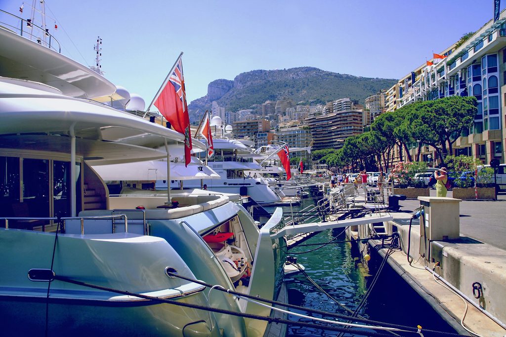 Motoryachten liegen achtern zur Hafenpromenade vertäut, im Hintergrund Hügel und Uferbauten