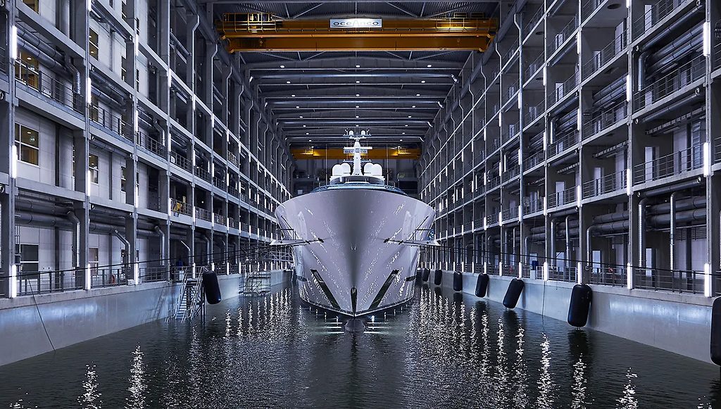 Prua di un superyacht in un bacino di cantiere navale al coperto, vista frontale tra strutture a più livelli