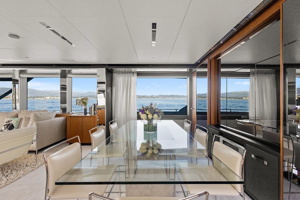 Table à manger en verre et coin salon sur le pont intérieur d'un yacht, avec vue sur le lac et les montagnes