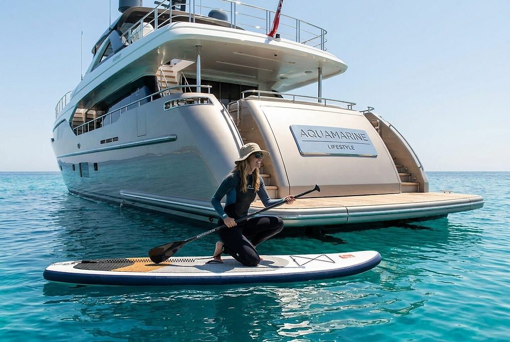 Woman kneeling on a paddleboard beside a superyacht's stern, anchored at sea