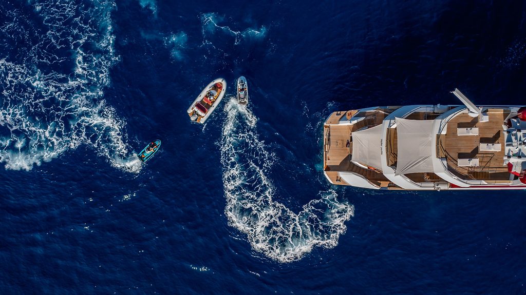 Aerial view of a motor yacht's stern with sun pads, small boats leaving wakes on dark blue sea