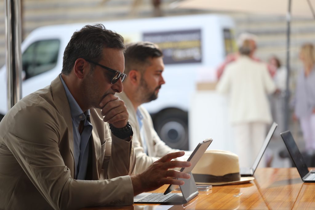 Deux hommes utilisant une tablette et des ordinateurs portables à une table en extérieur, camionnette et passants flous à l'arrière-plan