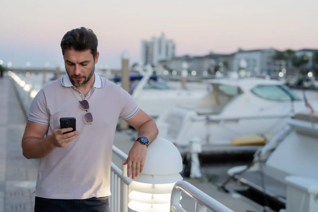 Homme consultant son téléphone sur un ponton de marina au crépuscule, yachts amarrés derrière lui