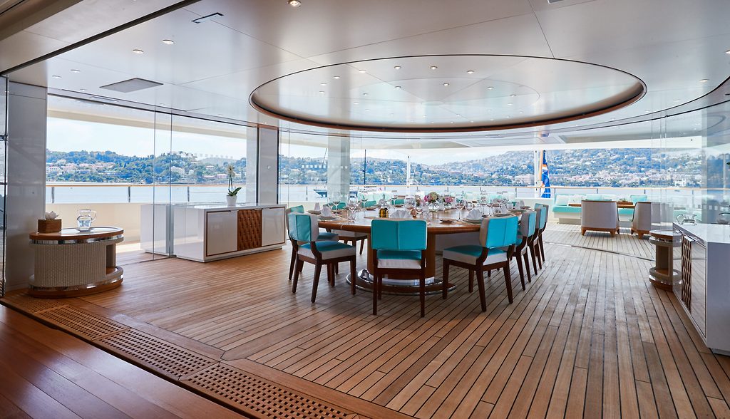 Essbereich auf dem Deck einer Superyacht, türkisfarbene Stühle um gedeckten Tisch, Blick auf Küstenbucht