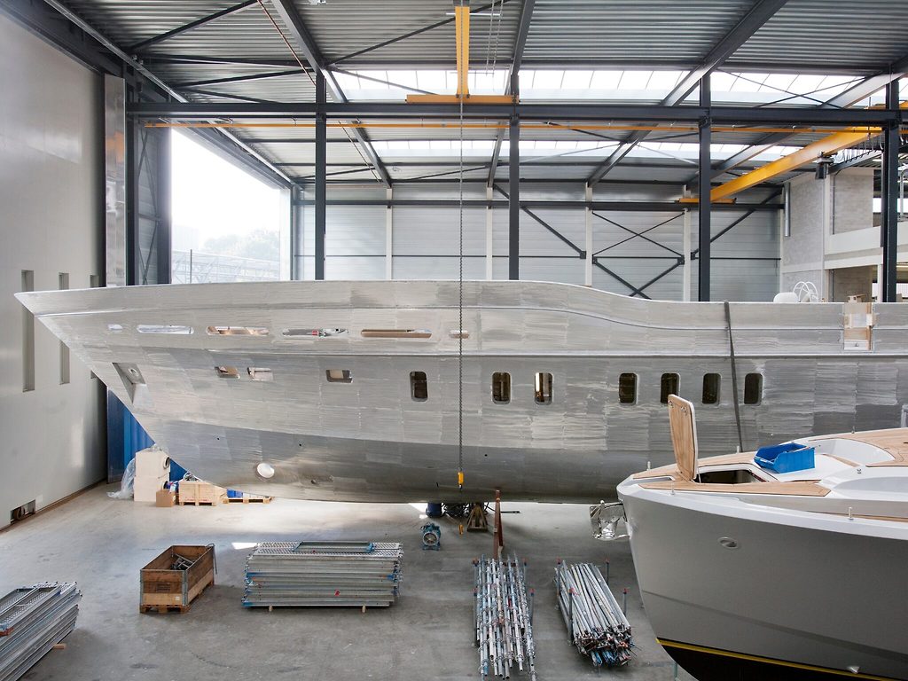 Unlackierter Aluminium-Yachtrumpf im Bau in einer Werfthalle, fertiger Rumpf daneben