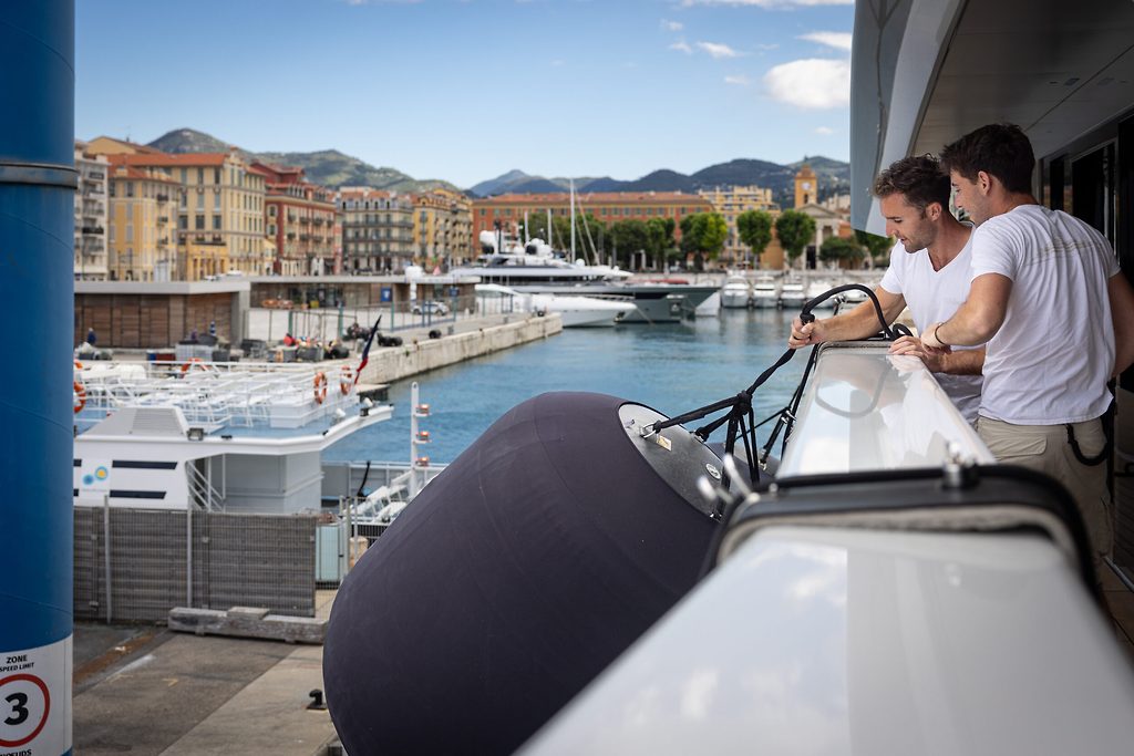 Deux membres d'équipage accoudés au bastingage d'un yacht avec vue sur un port de plaisance et des collines en arrière-plan