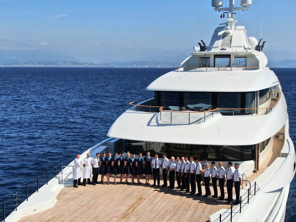 Uniformed crew lined up on the aft deck of a superyacht at sea, coastline in the distance