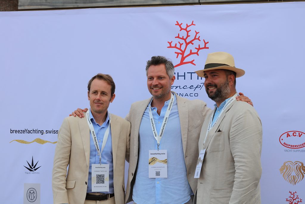 Tres hombres con acreditaciones de evento posando ante un panel de patrocinadores de un concepto yachting