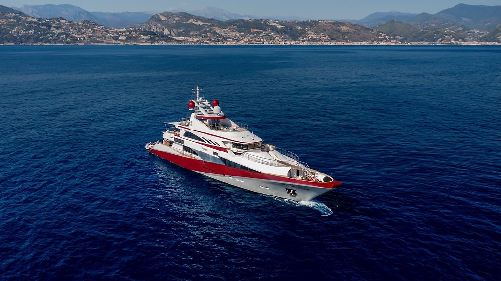 Motor yacht with red hull stripe cruising off a mountainous coastline