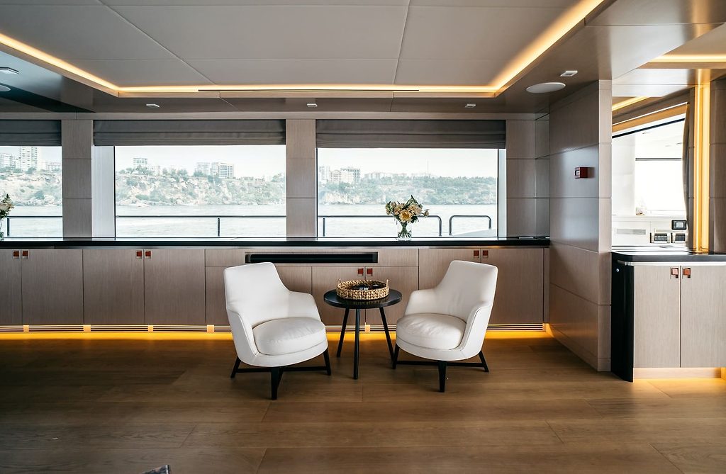 Lounge seating area with white armchairs on a yacht, coastal view through the windows