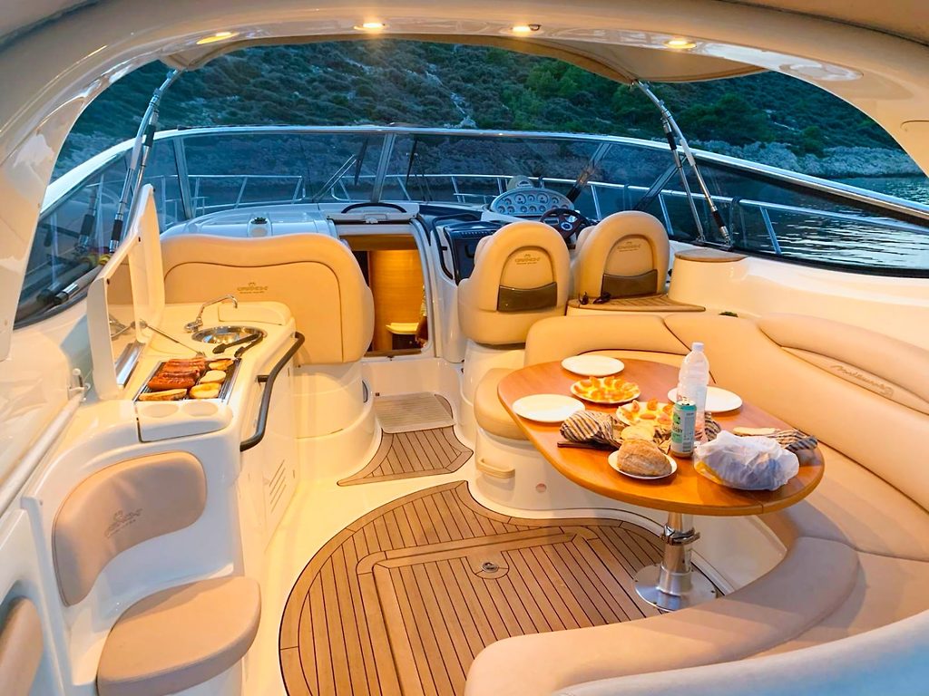 Cockpit of a motor yacht with dining table set for a meal and helm seats, near a coastline at dusk