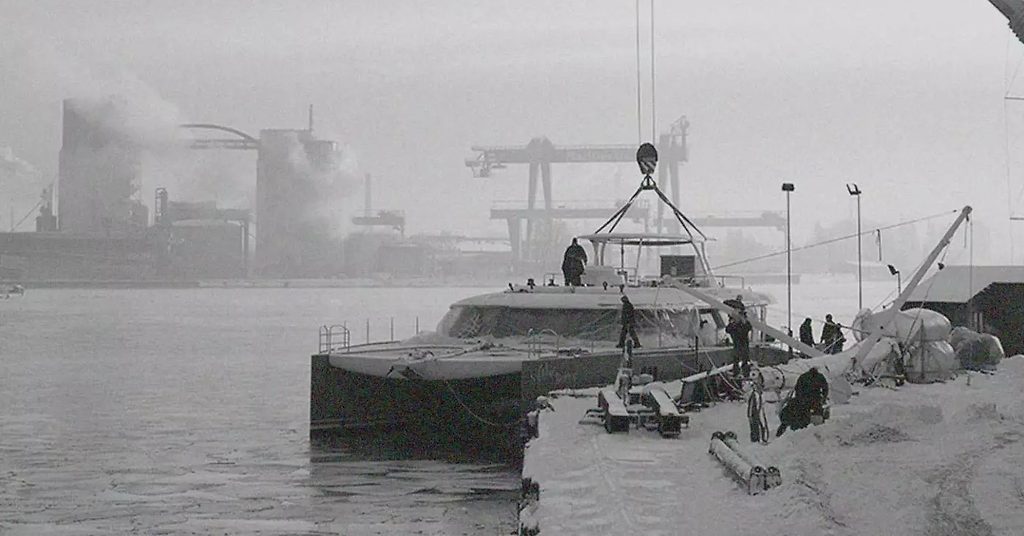 Catamaran hull being lifted by crane at a snowy shipyard dock