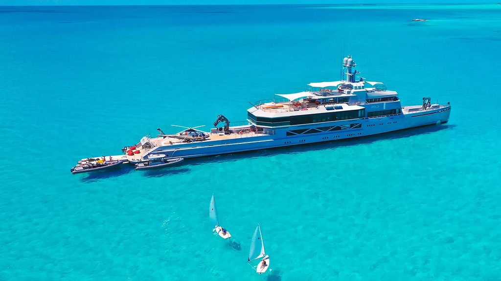 Yacht d'exploration au mouillage dans une eau turquoise avec un hélicoptère sur le pont et des annexes à couple, planchistes à proximité