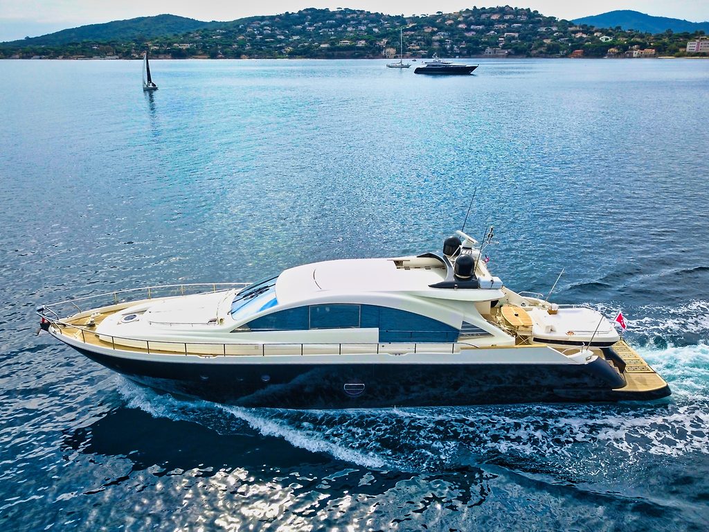 Yate a motor navegando cerca de una costa con colinas y barcos anclados