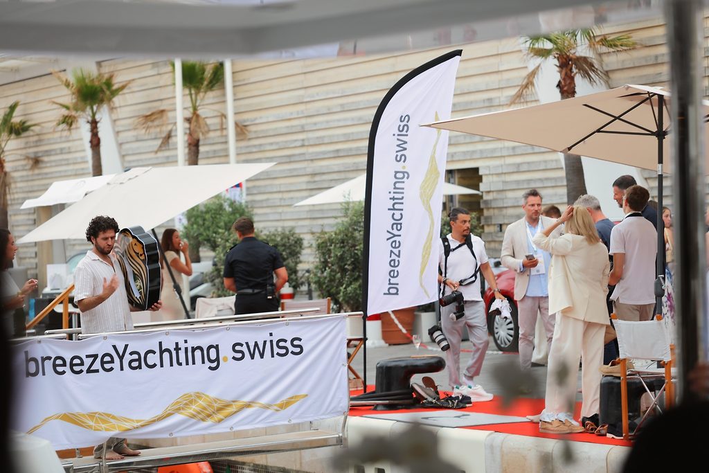 Гости на площадке с красной ковровой дорожкой и баннерами breezeYachting.swiss, пальмы на заднем плане