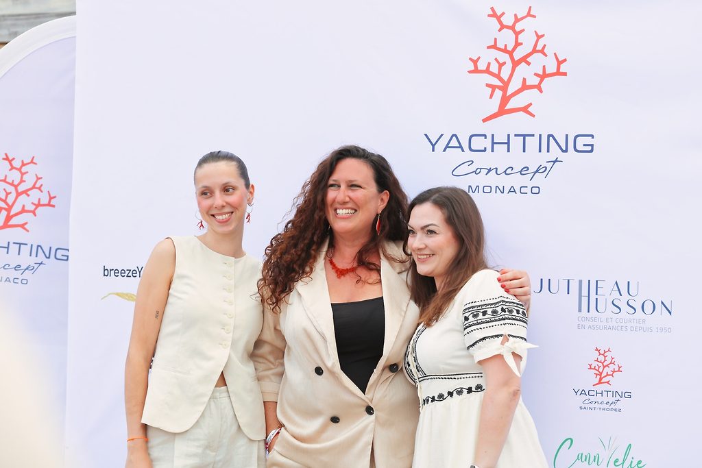 Три женщины позируют вместе на мероприятии Yachting Concept Monaco перед стендом спонсоров