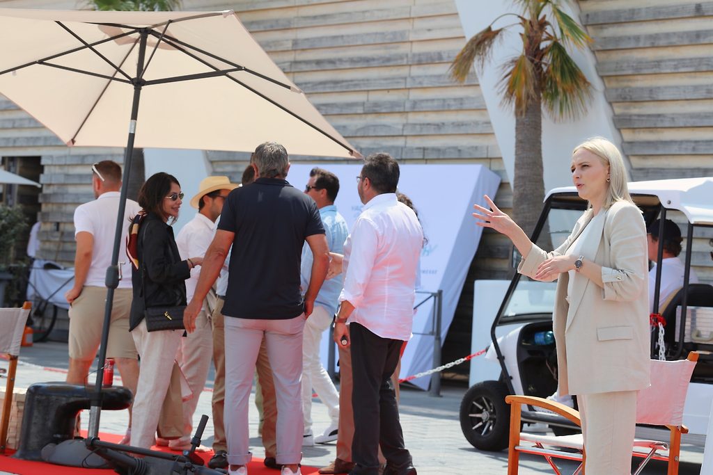 Женщина жестикулирует, разговаривая с группой людей под зонтом на открытом мероприятии в марине