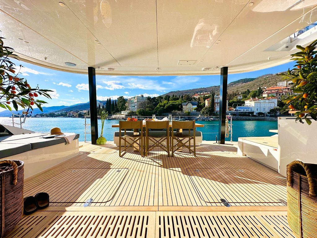 Cubierta de popa a la sombra con mesa de comedor en un catamarán, vista de una ciudad costera y una bahía