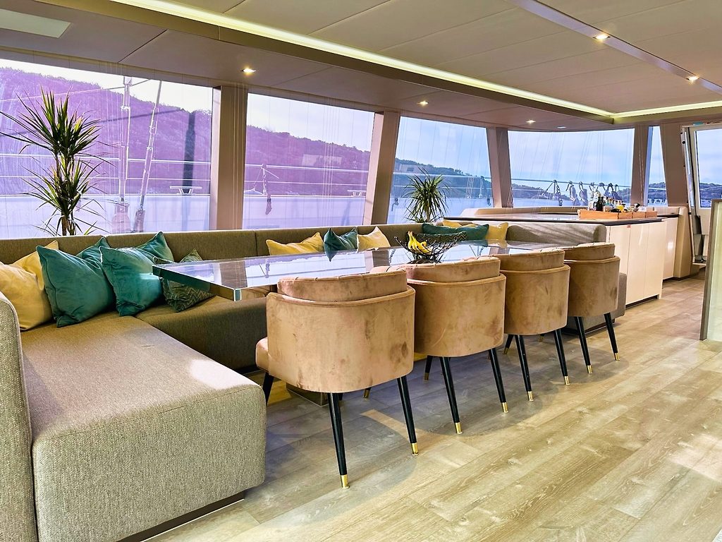 Salón con zona de estar y comedor a bordo de un catamarán, con ventanales panorámicos sobre un puerto