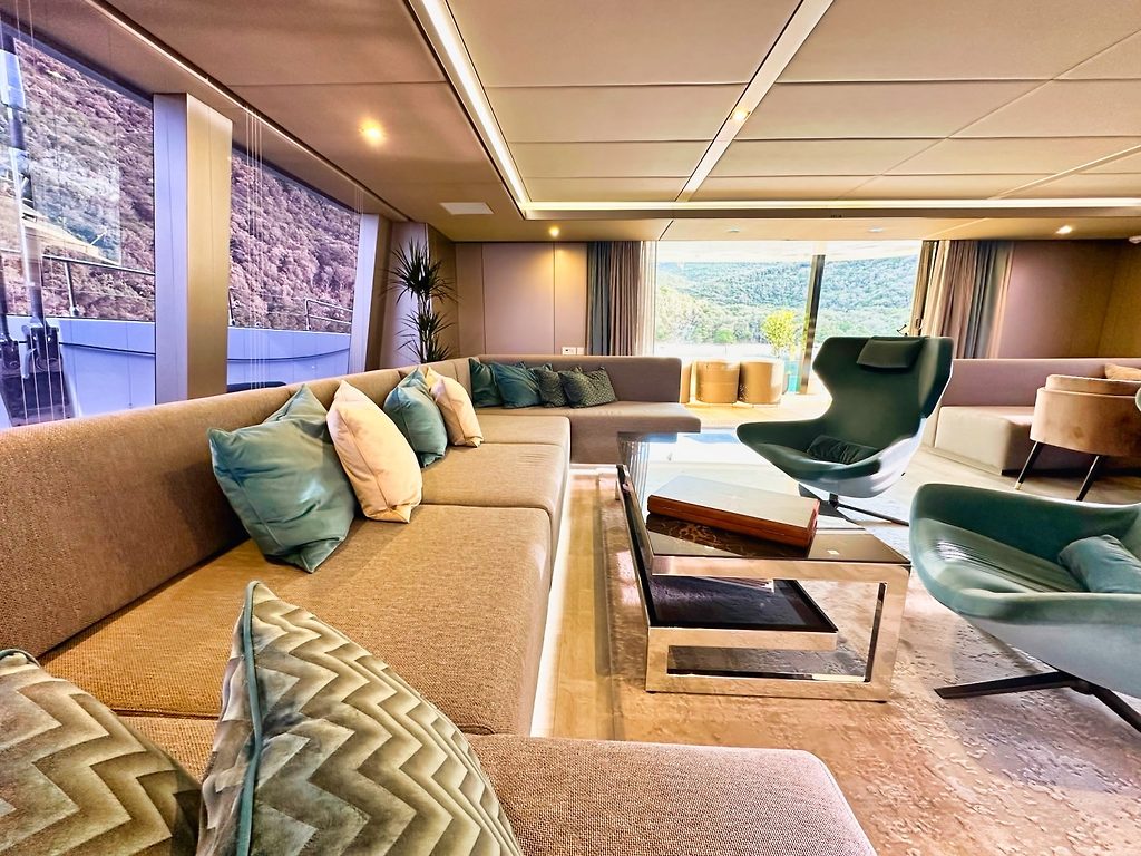 Salón de yate con sofá de esquina y sillones giratorios, grandes ventanales con vista a colinas