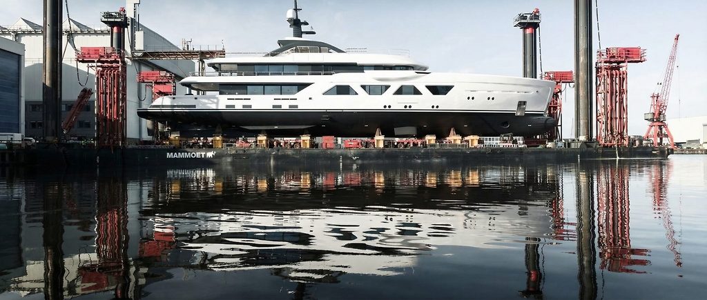 Yate a motor siendo lanzado desde un astillero entre grúas pórtico, casco reflejado en el agua
