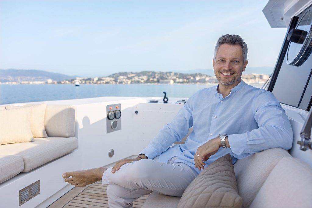 Mann sitzt im Lounge-Bereich auf dem Achterdeck einer Yacht, Küstenstadt im Hintergrund