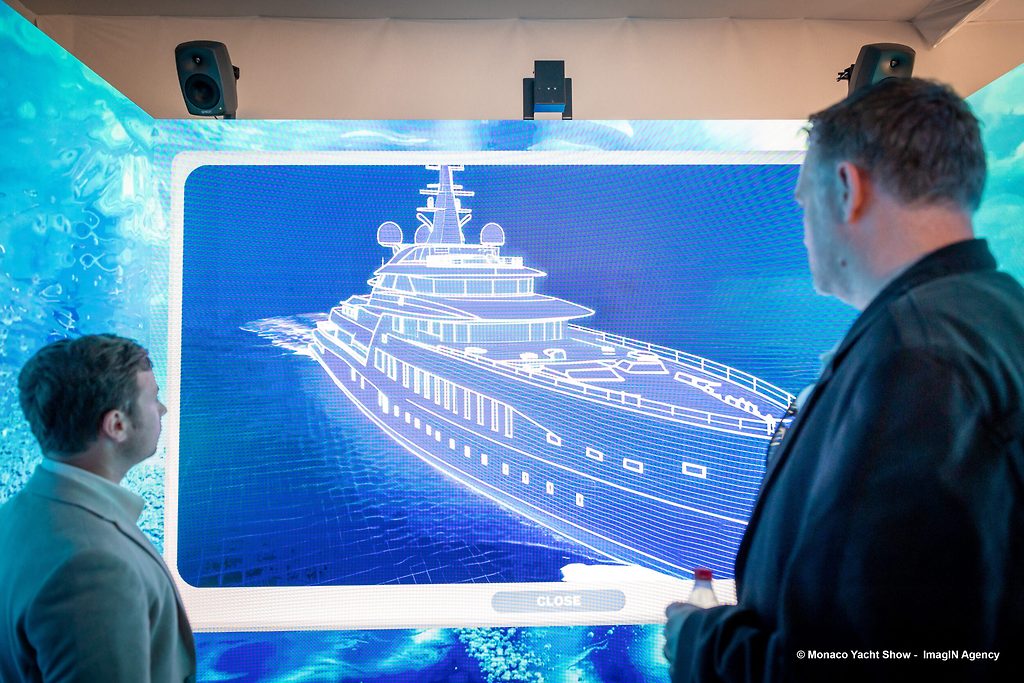 Two visitors viewing a superyacht wireframe rendering on a large digital screen at a boat show
