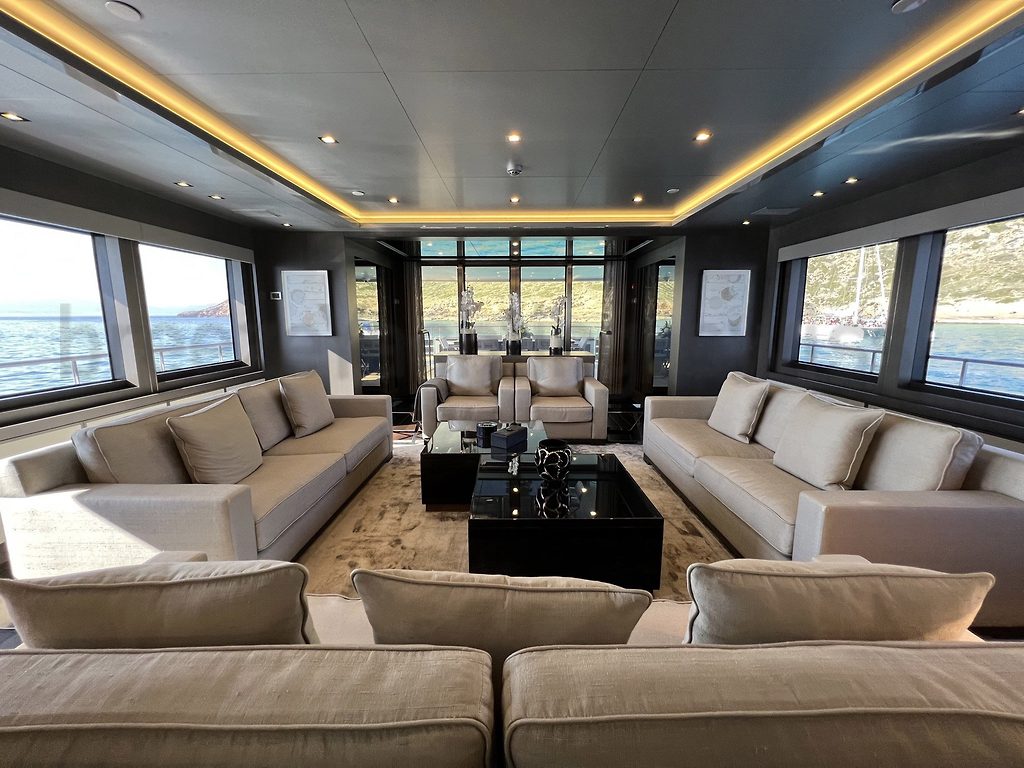 Salon d'un yacht à moteur avec canapés beiges et table basse, mouillage visible par les fenêtres